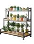 Grow24-Three-tier black metal plant stand with wood shelves and caster wheels holding multiple potted plants by a window